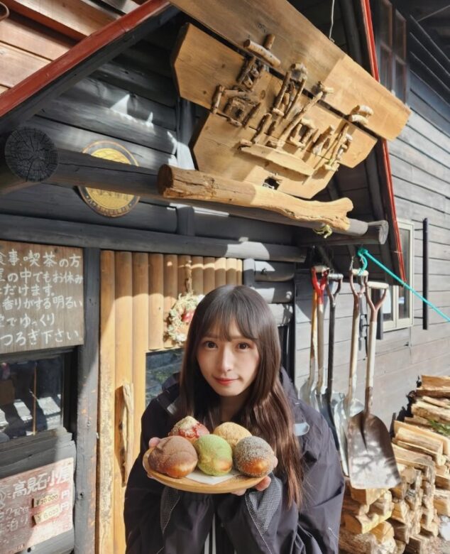 登山中に合ったパン屋の前でパンを持ってポーズをとる渡辺梨加さんの画像